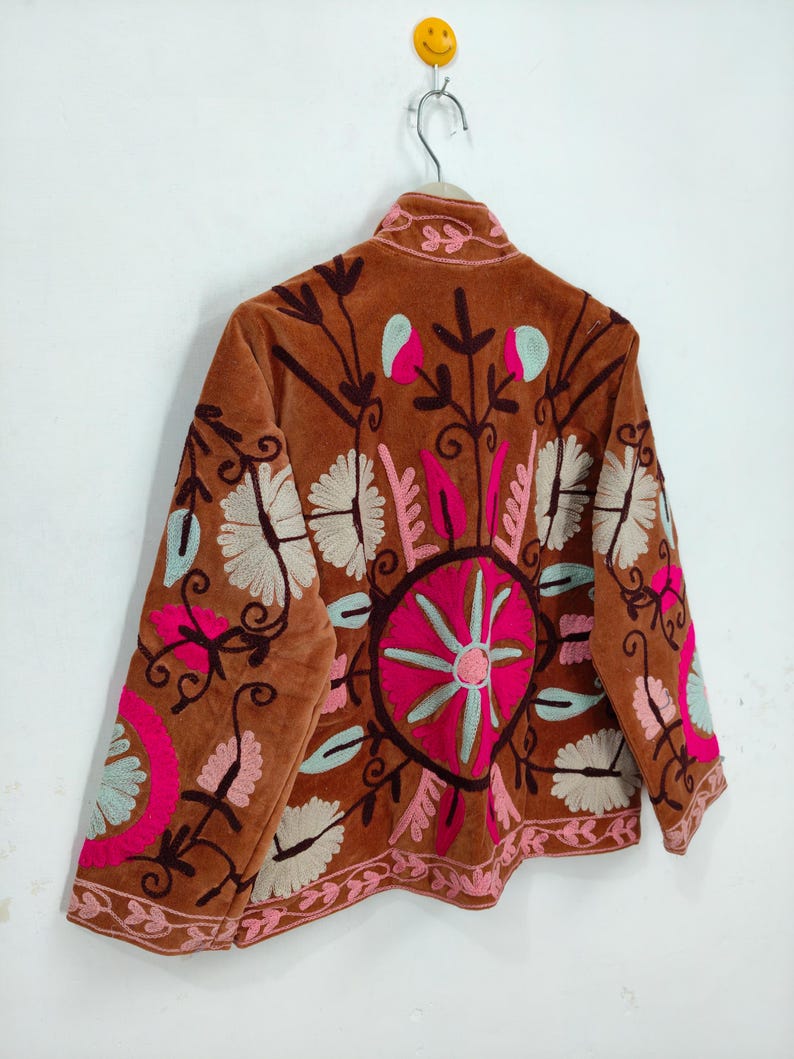 May include: A brown velvet jacket with intricate floral embroidery in pink, white, and teal. The jacket features a high collar and a detailed circular design on the back, showcasing a bohemian style.