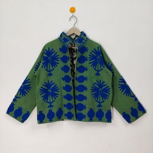 Könnte beinhalten: Eine grüne Jacke mit blau gestickten Blumenmustern. Die Jacke hat einen Stehkragen und lange Ärmel. Das Innenfutter hat ein schwarz-weißes Muster. Die Jacke hängt an einem Kleiderbügel.