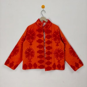 May include: An orange jacket with a mandarin collar and long sleeves. The jacket features a repeating pattern of red floral and circular designs. The jacket is hanging on a hanger against a white background.