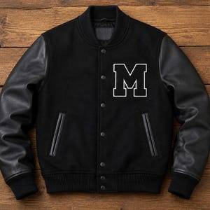 Könnte beinhalten: Eine schwarze Collegejacke mit einem Körper aus Wollmischgewebe und Ärmeln aus Leder. Die Jacke hat einen gerippten Kragen, Bündchen und Saum, eine Druckknopfleiste, zwei Seitentaschen mit Lederbesatz und einen großen weißen Buchstaben M auf der Brust.