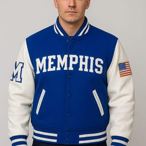Pode incluir: Casaco universitário azul e branco com a palavra "MEMPHIS" no peito. O casaco tem mangas brancas, um pequeno patch da bandeira americana e punhos e gola listrados. A letra "M" está na manga esquerda.