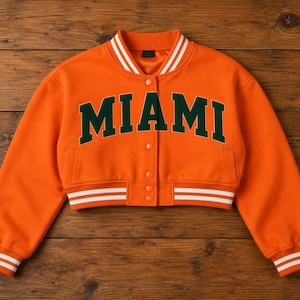 May include: Orange varsity jacket with the word "MIAMI" in dark green block letters across the chest. The jacket has white stripes on the collar, cuffs, and hem. It features a button-up front and two pockets.