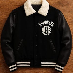 Puede incluir: Chaqueta universitaria negra con texto "Brooklyn" blanco y un logotipo de baloncesto. La chaqueta tiene un cuello de piel sintética blanco, mangas de cuero negro y puños y dobladillo a rayas blancas. Diseño deportivo clásico.