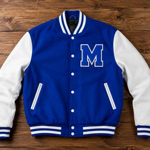 Può includere: Una giacca varsity blu reale con maniche bianche, caratterizzata da una grande lettera M bianca sul petto sinistro. La giacca presenta una chiusura frontale con bottoni a pressione, due tasche laterali con bordino bianco e polsini e colletto a coste a righe blu e bianche.