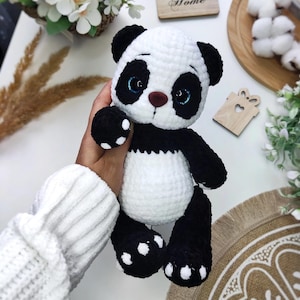 May include: A handmade crochet panda bear in black and white, with blue eyes and a brown nose. The panda has black paws with white spots. The bear is a soft toy.