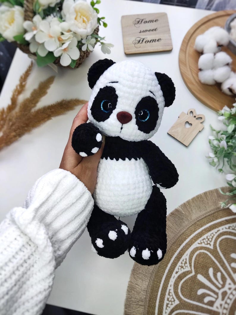May include: A handmade crochet panda bear in black and white, with blue eyes and a brown nose. The panda has black paws with white spots. The bear is a soft toy.