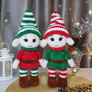 May include: Two handmade crochet elf dolls. One elf wears a red outfit with green and white striped legs and a red and white striped hat. The other elf wears a green outfit with green and white striped legs and a green and white striped hat. Both dolls have white faces and brown shoes.