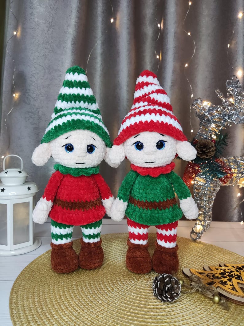 May include: Two handmade crochet elf dolls. One elf wears a red outfit with green and white striped legs and a red and white striped hat. The other elf wears a green outfit with green and white striped legs and a green and white striped hat. Both dolls have white faces and brown shoes.