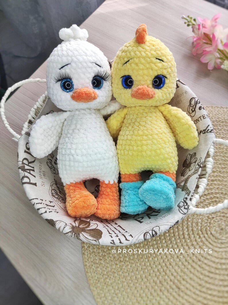 May include: Two handmade crochet duck toys. One is white with orange feet and a white tuft, the other is yellow with blue booties and an orange crest. Both have blue eyes and orange beaks, sitting in a decorative basket.