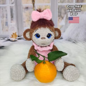 Könnte beinhalten: Eine handgefertigte Häkel-Affenpuppe mit einer rosa Schleife, Kragen und großen blauen Augen. Der Affe hält eine frische Orange mit grünen Blättern. Der Text "Crochet pattern PDF" und eine US-Flagge befinden sich in der oberen rechten Ecke.