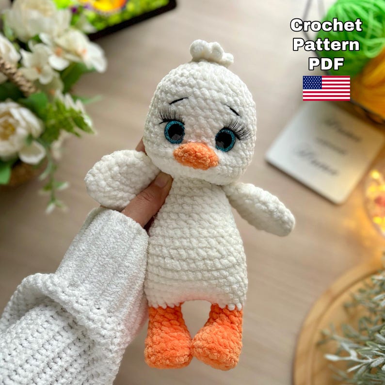 May include: A handmade white crochet duck with orange feet and beak, and blue eyes with long eyelashes. The duck is being held by a person. Text on the image reads "Crochet Pattern PDF" with a US flag.