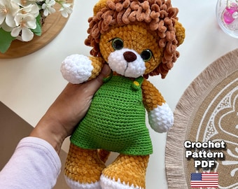 Può includere: Un leone di peluche all'uncinetto con criniera marrone, corpo arancione e dettagli bianchi. Indossa una tuta verde. L'immagine include il testo "Crochet Pattern PDF" e una bandiera americana.
