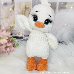 May include: A handmade white crochet duck with orange feet and beak, and large blue eyes with long eyelashes. The duck is waving one wing. The text "@PROSKURYAKOVA_KNITS" is at the bottom.