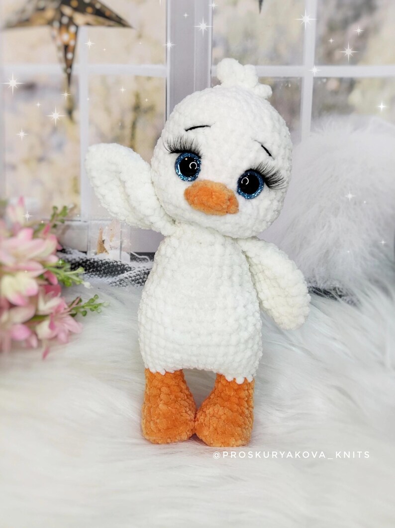 May include: A handmade white crochet duck with orange feet and beak, and large blue eyes with long eyelashes. The duck is waving one wing. The text "@PROSKURYAKOVA_KNITS" is at the bottom.