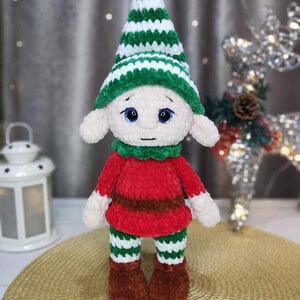 May include: A handmade plush elf doll with a green and white striped hat, green collar, red shirt, and green and white striped pants. The elf has brown shoes and white hands. The doll has blue eyes and is standing on a gold surface.