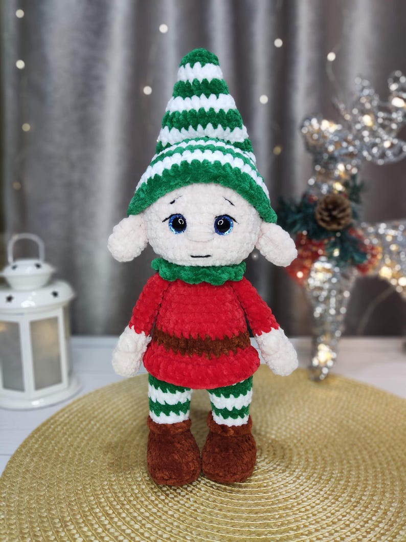 May include: A handmade plush elf doll with a green and white striped hat, green collar, red shirt, and green and white striped pants. The elf has brown shoes and white hands. The doll has blue eyes and is standing on a gold surface.