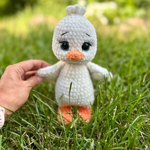 May include: A handmade white crochet duckling with an orange beak and feet, and large blue eyes with long eyelashes. The duckling has small wings and is set against a background of green grass.