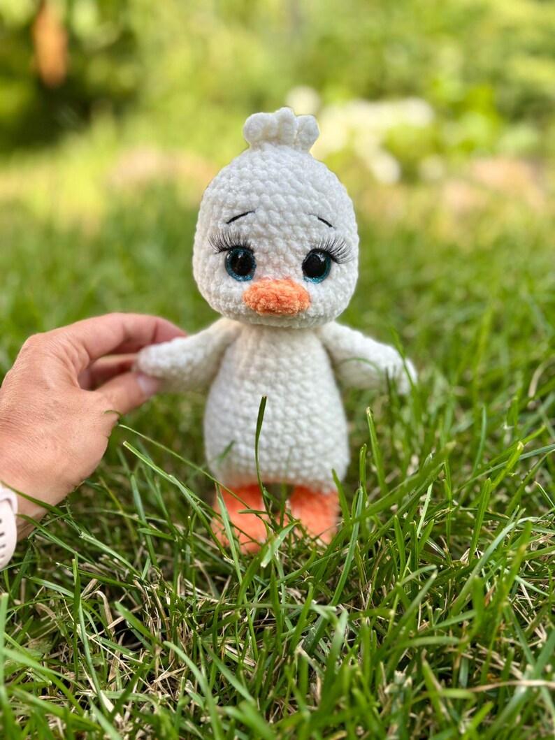 May include: A handmade white crochet duckling with an orange beak and feet, and large blue eyes with long eyelashes. The duckling has small wings and is set against a background of green grass.