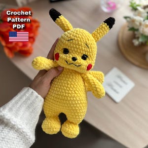 Peut inclure: Peluche Pikachu jaune au crochet, faite à la main, avec des yeux noirs, des joues rouges et des oreilles à pointes noires. La peluche a de petits bras et jambes. L'image comprend le texte "Crochet Pattern PDF" et un drapeau américain.