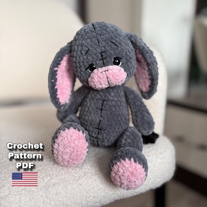 Puede incluir: Un peluche de conejo de ganchillo gris hecho a mano con detalles rosas en las orejas, la nariz y los pies. El peluche está sentado en una silla blanca. La imagen incluye el texto "Crochet Pattern PDF" y la bandera estadounidense.