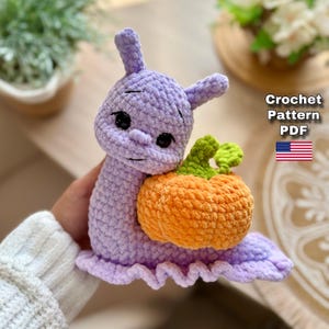 May include: A handmade crochet snail in lavender with a pumpkin. The snail has black eyes and a smiling face. The pumpkin is orange with green leaves. The text "Crochet Pattern PDF" is visible, along with the American flag.