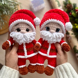 Puede incluir: Dos mu&ntilde;ecos de Pap&aacute; Noel hechos a mano con ganchillo. Cada mu&ntilde;eco tiene un traje rojo con ribetes blancos, una barba blanca y un gorro rojo con un pomp&oacute;n blanco. Los mu&ntilde;ecos se sostienen delante de un fondo borroso.