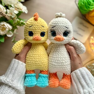 May include: Two handmade crochet duck dolls. One is yellow with blue boots and an orange beak and crest. The other is white with orange feet. Both have large blue eyes and are being held by a person wearing a white sweater.