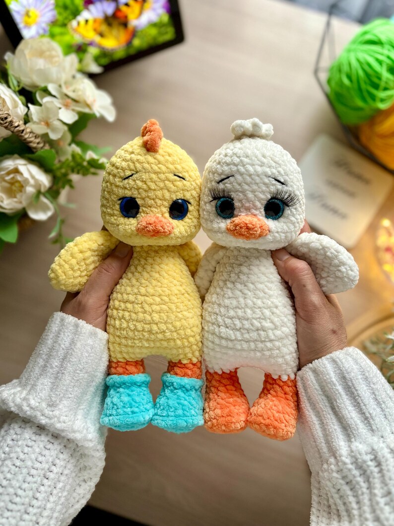 May include: Two handmade crochet duck dolls. One is yellow with blue boots and an orange beak and crest. The other is white with orange feet. Both have large blue eyes and are being held by a person wearing a white sweater.