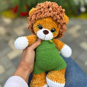 May include: A handmade crochet lion plush toy with a brown mane, orange body, and white accents on the paws. The lion wears a green dress. The toy is soft and cuddly, perfect for children.