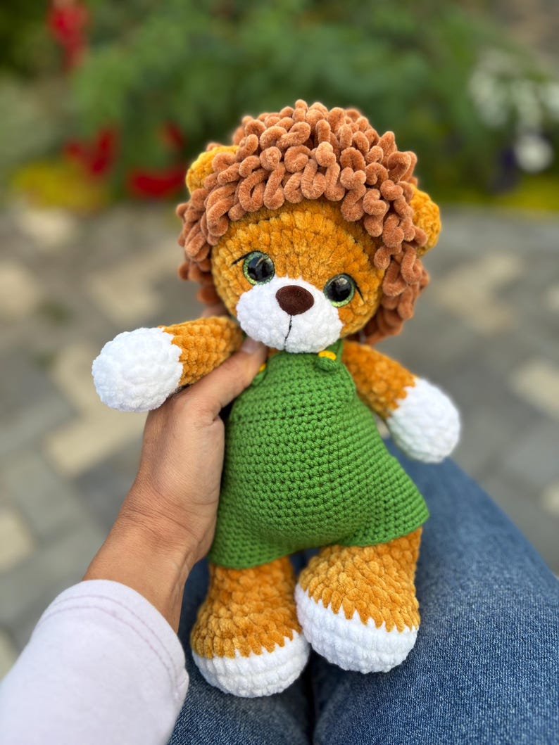May include: A handmade crochet lion plush toy with a brown mane, orange body, and white accents on the paws. The lion wears a green dress. The toy is soft and cuddly, perfect for children.