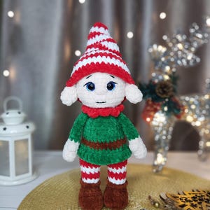 May include: A handmade crochet elf doll with a red and white striped hat, green tunic, and red and white striped pants. The doll has white ears, blue eyes, and brown boots. The elf is a festive holiday decoration.