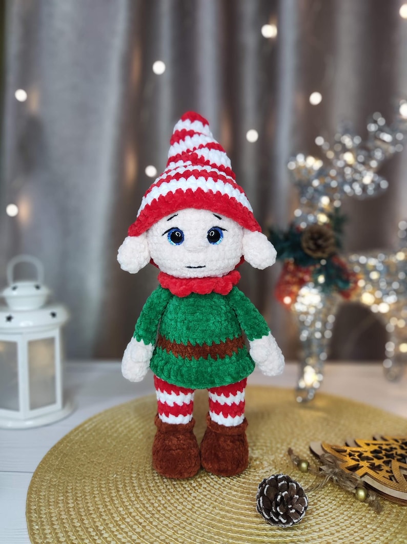 May include: A handmade crochet elf doll with a red and white striped hat, green tunic, and red and white striped pants. The doll has white ears, blue eyes, and brown boots. The elf is a festive holiday decoration.