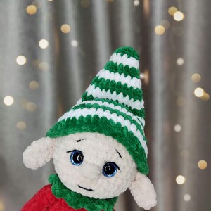 May include: A handmade plush elf doll with a red body, green collar, and a green and white striped hat. The doll has large blue eyes and small, pointed ears. The background features soft, blurred lights.