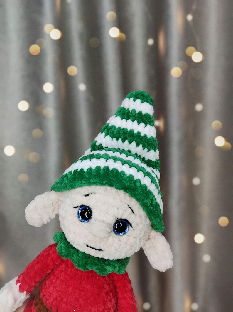 May include: A handmade plush elf doll with a red body, green collar, and a green and white striped hat. The doll has large blue eyes and small, pointed ears. The background features soft, blurred lights.