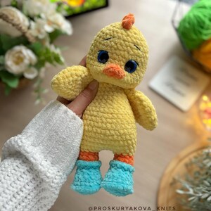 May include: A handmade yellow crochet chick with orange feet and a small orange crest. The chick has blue eyes and is wearing blue boots. The text "@PROSKURYAKOVA_KNITS" is at the bottom of the image.