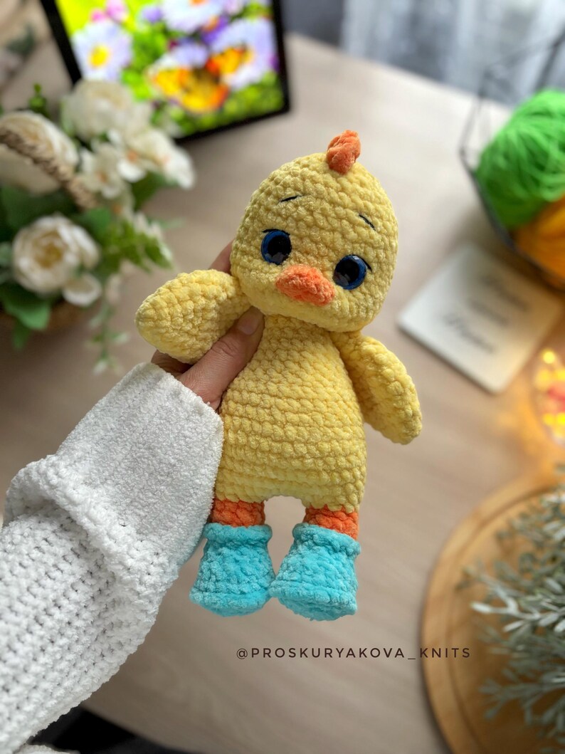May include: A handmade yellow crochet chick with orange feet and a small orange crest. The chick has blue eyes and is wearing blue boots. The text "@PROSKURYAKOVA_KNITS" is at the bottom of the image.