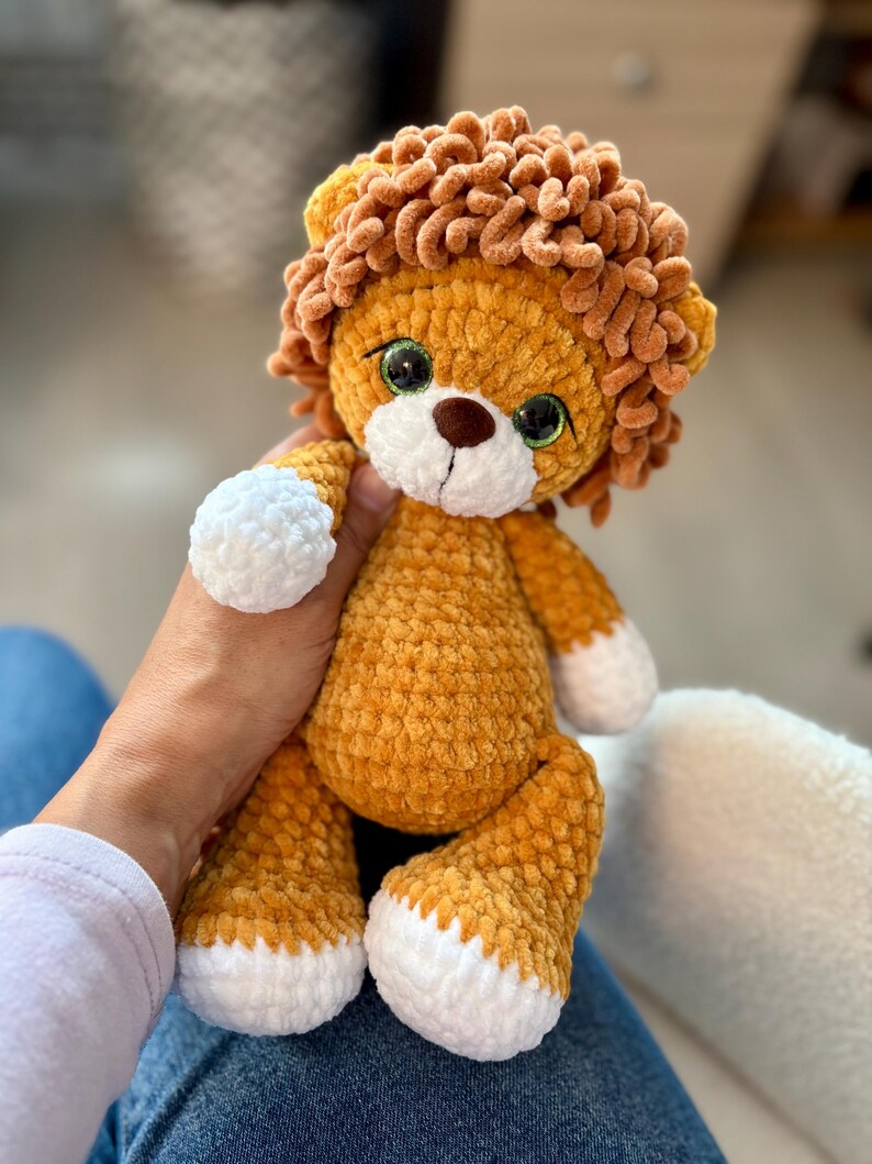 May include: A handmade, plush lion toy with a golden-yellow body and a brown mane. The lion has white paws and a white face, with green eyes and a brown nose. The toy is soft and cuddly, perfect for children.