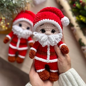Puede incluir: Dos mu&ntilde;ecos de Pap&aacute; Noel hechos a mano con ganchillo. Cada mu&ntilde;eco tiene un traje rojo, gorro y botas y guantes marrones. Los mu&ntilde;ecos tienen barbas blancas y caras sonrientes. Uno est&aacute; enfocado, el otro ligeramente borroso.