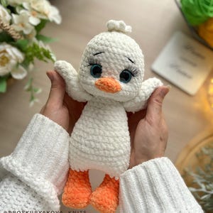 May include: A handmade white crochet duck toy with orange feet and beak, and blue eyes with long eyelashes. The duck has outstretched wings and a small tuft of white yarn on its head. The toy is held in front of a soft, blurred background.