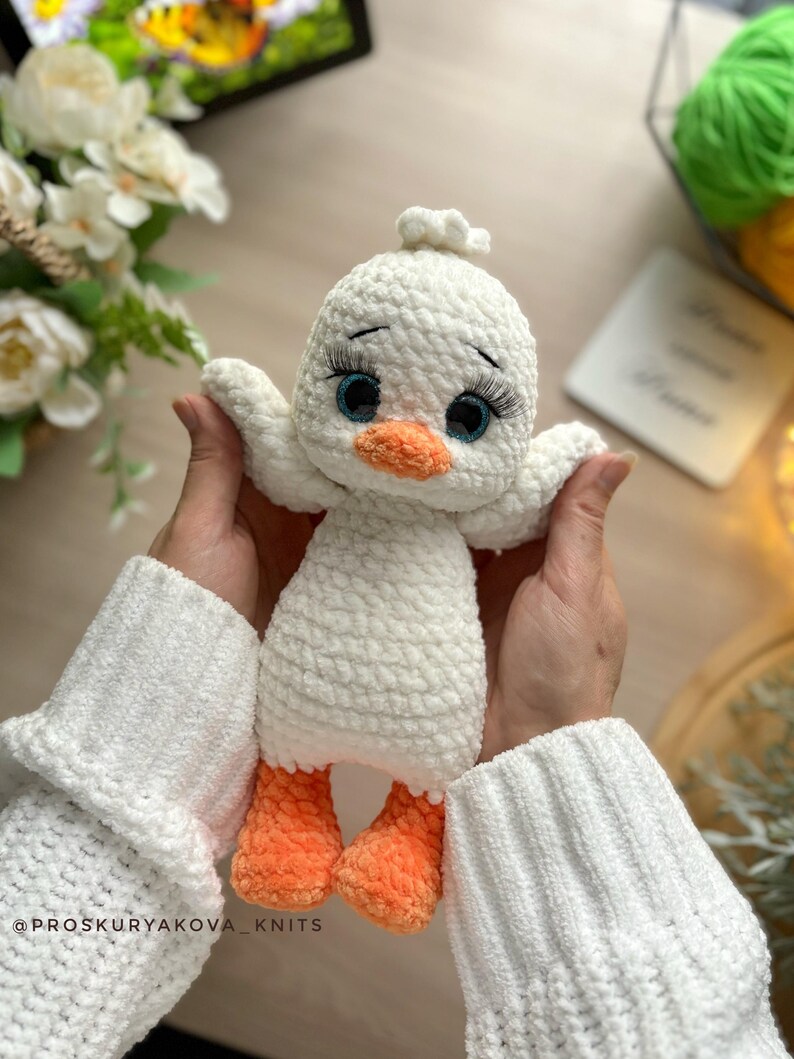 May include: A handmade white crochet duck toy with orange feet and beak, and blue eyes with long eyelashes. The duck has outstretched wings and a small tuft of white yarn on its head. The toy is held in front of a soft, blurred background.