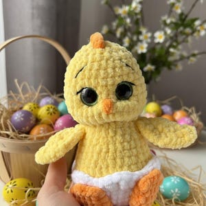 Pode incluir: Um pintinho de croch&ecirc; amarelo feito &agrave; m&atilde;o com olhos grandes pretos, bico e p&eacute;s laranja. O pintinho est&aacute; usando uma fralda branca. Ovos de P&aacute;scoa em uma cesta e em uma mesa ao fundo.