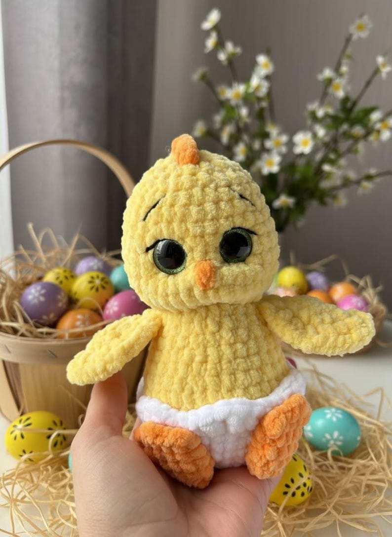 Pode incluir: Um pintinho de croch&ecirc; amarelo feito &agrave; m&atilde;o com olhos grandes pretos, bico e p&eacute;s laranja. O pintinho est&aacute; usando uma fralda branca. Ovos de P&aacute;scoa em uma cesta e em uma mesa ao fundo.
