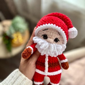 Puede incluir: Una mu&ntilde;eca de ganchillo hecha a mano de Pap&aacute; Noel. La mu&ntilde;eca tiene un gorro rojo con ribete blanco, una barba blanca y un traje rojo con detalles blancos. La cara de la mu&ntilde;eca es beige con ojos negros y una nariz peque&ntilde;a.