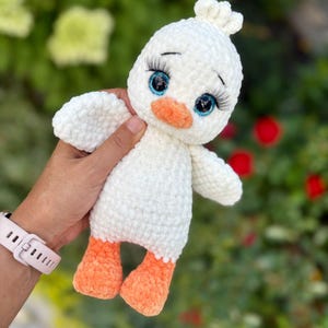May include: A handmade white crochet duckling plush toy with blue eyes, long eyelashes, and an orange beak and feet. The duckling has small wings and a tuft of white yarn on its head. The toy is held against a blurred green and red background.