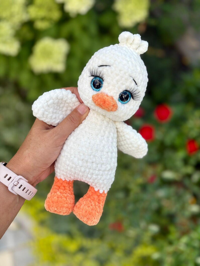 May include: A handmade white crochet duckling plush toy with blue eyes, long eyelashes, and an orange beak and feet. The duckling has small wings and a tuft of white yarn on its head. The toy is held against a blurred green and red background.