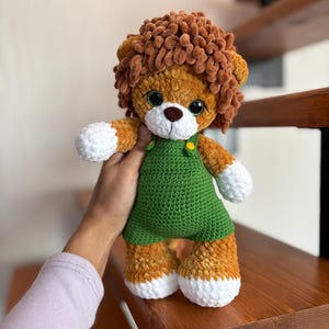 May include: A handmade crochet lion plush toy. The lion has a brown mane, a white muzzle, and large black eyes. It wears a green overall dress with yellow button details. The lion's paws and feet are white.