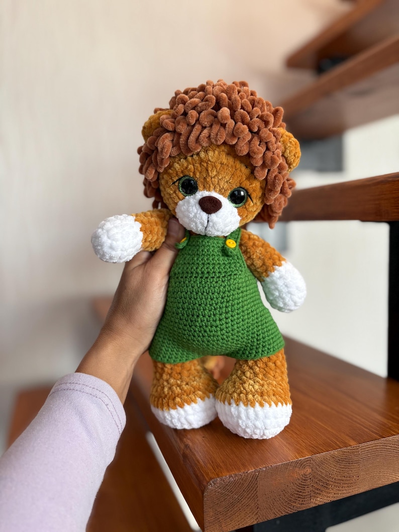 May include: A handmade crochet lion plush toy. The lion has a brown mane, a white muzzle, and large black eyes. It wears a green overall dress with yellow button details. The lion's paws and feet are white.