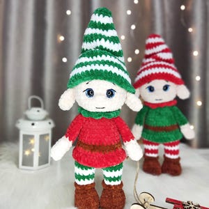 May include: Two handmade crochet elf dolls. One elf wears a green and white striped hat, red shirt, brown belt, green and white striped pants, and brown boots. The other elf wears a red and white striped hat, green shirt, and red and white striped pants. A small wooden sled is in the foreground.