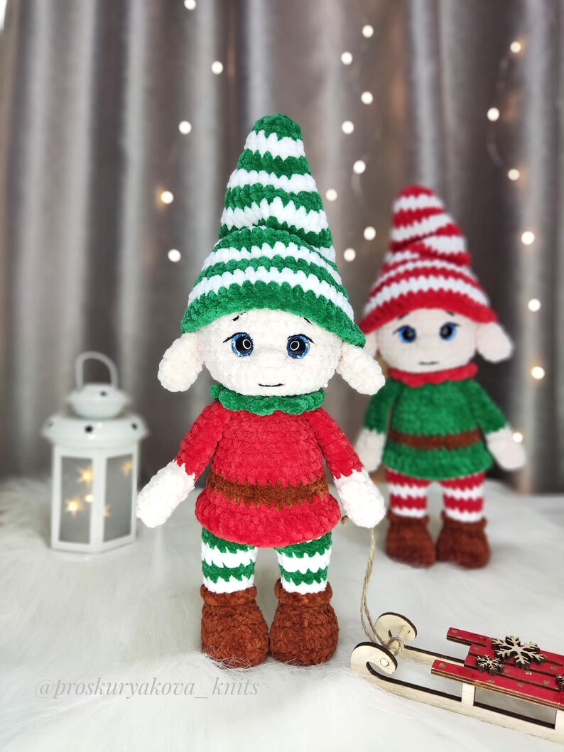 May include: Two handmade crochet elf dolls. One elf wears a green and white striped hat, red shirt, brown belt, green and white striped pants, and brown boots. The other elf wears a red and white striped hat, green shirt, and red and white striped pants. A small wooden sled is in the foreground.