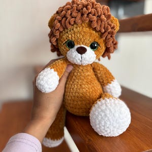 May include: A handmade crochet lion plush toy. The lion has a brown mane, a tan body, and white accents on its paws and muzzle. It has large, expressive eyes and a cute, friendly expression. The plush is soft and cuddly.
